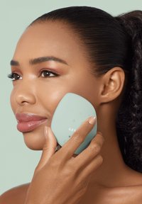 Dispositif facial aquamarine avec boutons, tenu contre une joue. Forme lisse et ergonomique. Le modèle a les lèvres brillantes et un vernis à ongles neutre.