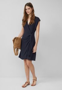 Marineblaues Wickelkleid mit kurzen Ärmeln und einer seitlichen Binde-Detail. Kombiniert mit flachen braunen Sandalen und einer gewebten hellbraunen Tasche. Glatte Stoffstruktur.