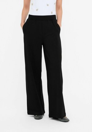 Kobieta w czarnych szerokich spodniach z elastycznym pasem, z rękami w kieszeniach, w połączeniu z wzorzystymi płaskimi butami.