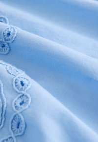 Tessuto azzurro chiaro con una texture morbida, caratterizzato da un ricamo intricato lungo un bordo, con motivi circolari realizzati con filo dello stesso colore.