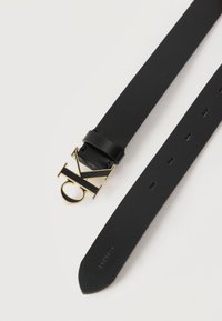 Μαύρη δερμάτινη ζώνη με χρυσή αγκράφα λογότυπο "CK" της Calvin Klein και πολλαπλές τρύπες ρύθμισης, σε λευκό φόντο.