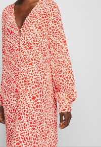 Červené a bílé vzorované šaty s výstřihem do V, dlouhými rukávy a volným střihem. Obsahují složité abstraktní tvary a přední zip.