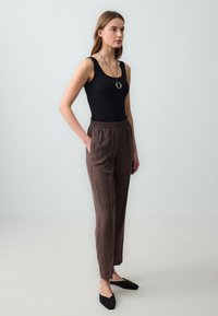 Zwarte geribbelde tanktop, bruine broek met elastische taille, en zwarte platte schoenen. Model staat met één hand in de zak, van natuurlijke stoffen en met strakke lijnen.