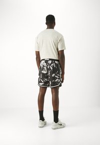 Conjunto deportivo masculino que incluye una camiseta de color beige claro con un estampado, pantalones cortos negros con gráficos abstractos en blanco, calcetines negros y zapatillas blancas.