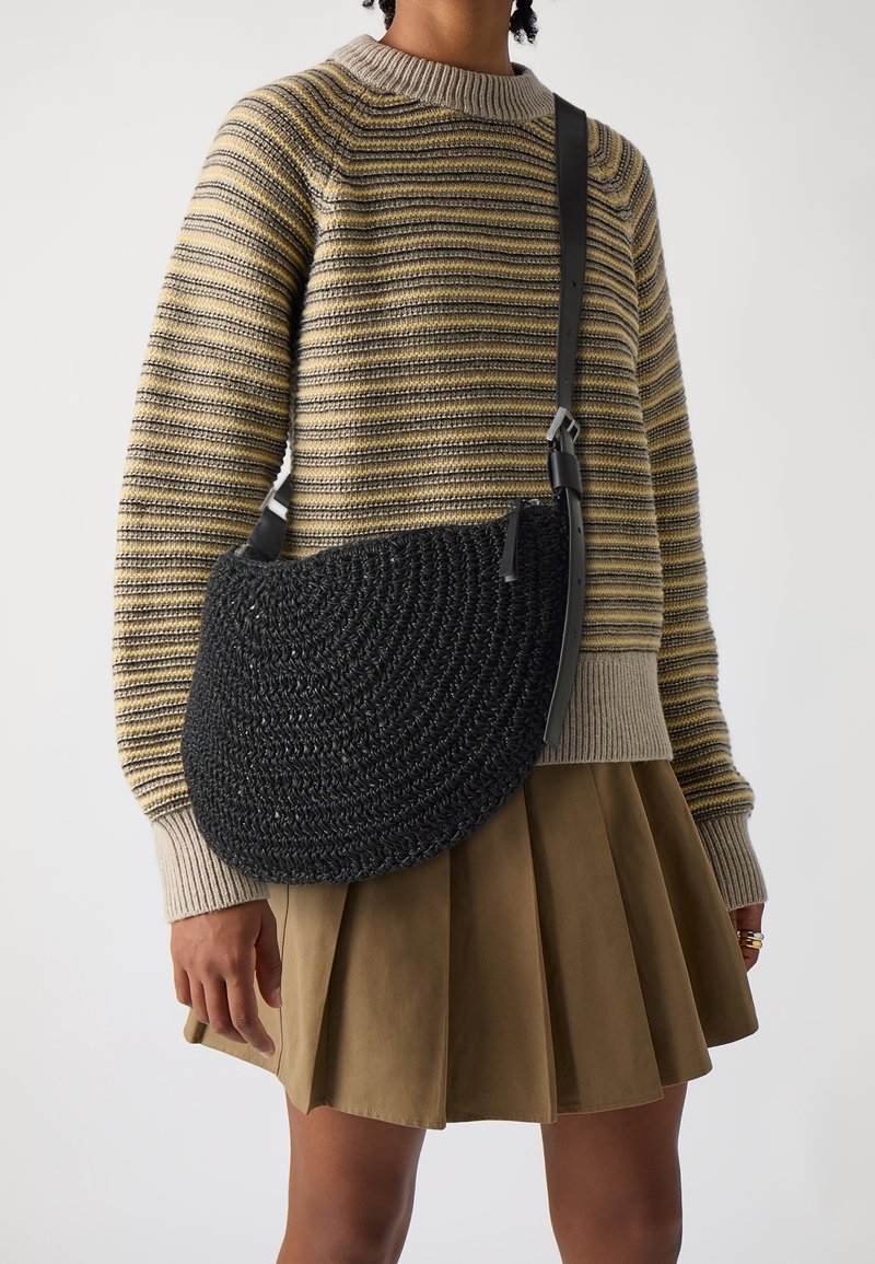 Personne portant un pull rayé beige et gris, une jupe plissée marron, et portant un sac bandoulière tressé noir avec une sangle en cuir.
