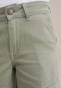 Kaki šorti ar struktūru audumu, priekšējo kabatu, jostas cilpiņām un metāla pogu akcentu ar logo. Jostasvieta ir nedaudz pacelta.