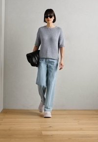 Grijze gebreide trui met korte mouwen, lichtblauwe wijde jeans en witte platform sneakers. Zwarte handtas en zonnebril. Houten vloer als achtergrond.