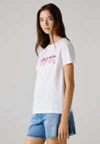 Νεαρή γυναίκα με μακριά καστανά μαλλιά, φορώντας λευκό t-shirt Red Bull Racing και ανοιχτόχρωμα τζιν σορτς μπροστά σε απλό φόντο.