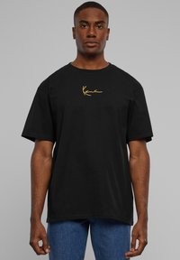 Black cotton t-shirt with short sleeves and a rounded neckline, featuring a gold embroidered logo on the left chest. Worn with blue denim jeans.