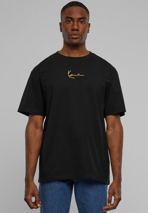 Sort bomulds T-shirt med korte ærmer og rund halsudskæring, prydet med et guld broderet logo på venstre bryst. Båret med blå denim jeans.