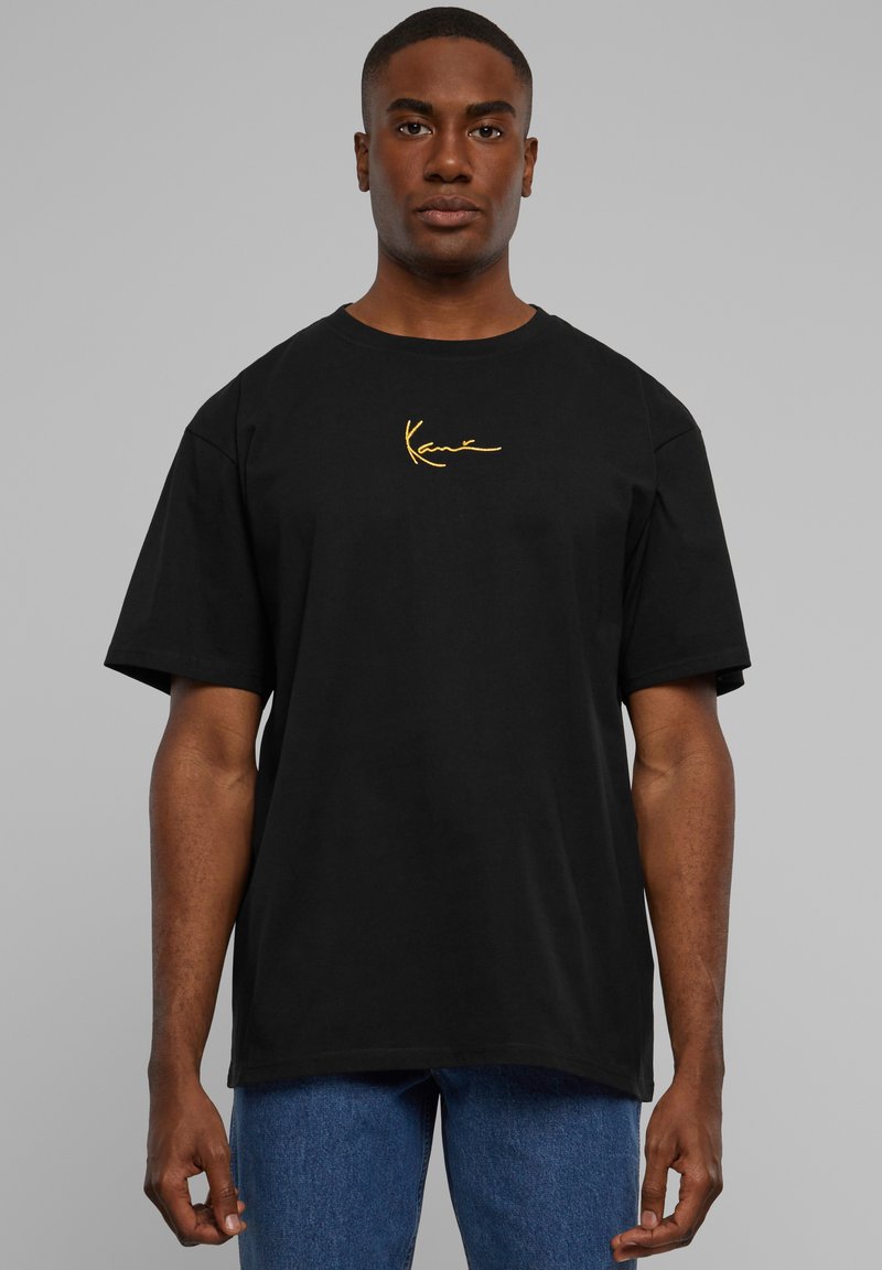 Black cotton t-shirt with short sleeves and a rounded neckline, featuring a gold embroidered logo on the left chest. Worn with blue denim jeans.