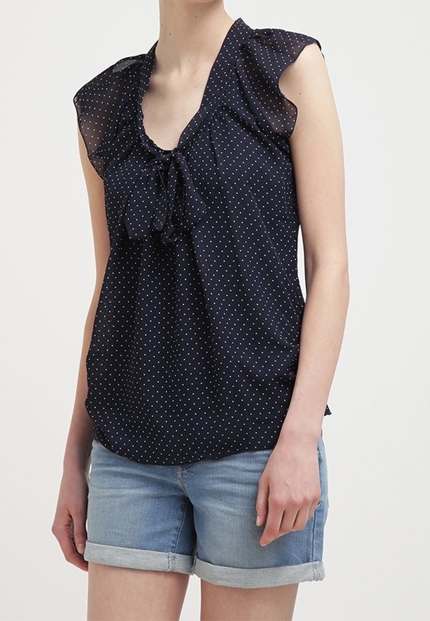 Femme portant un chemisier sans manches bleu marine à pois blancs avec des épaules volantées, associé à un short en denim retroussé.