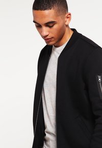 Giacca bomber nera con chiusura frontale a zip, colletto a costine e tasche laterali, con una tasca con zip sulla manica sinistra. Texture del tessuto morbida.