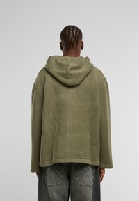 Pulover cu glugă de culoare verde măsliniu, cu croială lejeră, fabricat dintr-un material texturat. Prezintă mâneci lungi și o margine curbată ușor. Stilizat cu blugi gri.