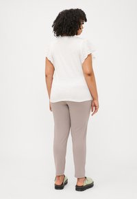 A woman with curly hair faces away, wearing a white shirt with decorative sleeves and beige pants, paired with green shoes.