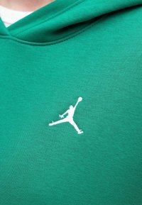 Zielona bluza z kapturem wykonana z miękkiego materiału, z białym haftowanym logo Jumpman po lewej stronie. Gładka faktura i luźny krój.