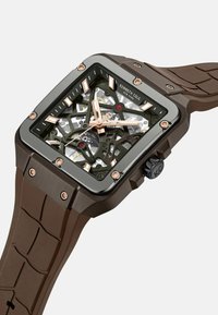 Vierkante skeleton horloge met een donkerbruine siliconen band, een grijs metalen frame, roségouden accenten en zichtbare ingewikkelde interne mechanismen.