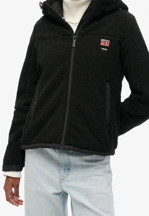 Veste polaire noire zippée avec capuche et petit logo rouge et blanc sur la poitrine, portée sur un pull blanc et un jean bleu clair.