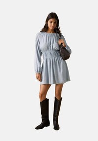 Hellblaues Kleid mit langen Ärmeln, geraffter Taille und ausgestelltem Rock. Kombiniert mit schwarzen kniehohen Stiefeln und einer kleinen dunklen Tasche.