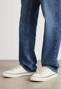 Baskets en toile blanches avec des lacets beiges et des accents noirs, associées à un jean en denim bleu à coupe droite et délavage léger.