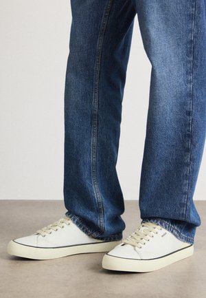 Weiße Canvas-Sneaker mit beigen Schnürsenkeln und schwarzen Akzenten, kombiniert mit blauen Jeans im geraden Schnitt und heller Waschung.