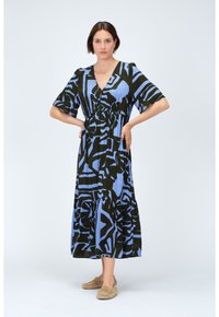 Robe maxi à motifs bleus et noirs avec un décolleté en V, des manches courtes et une jupe à étages, fabriquée en tissu léger. Associée à des chaussures à enfiler beiges.
