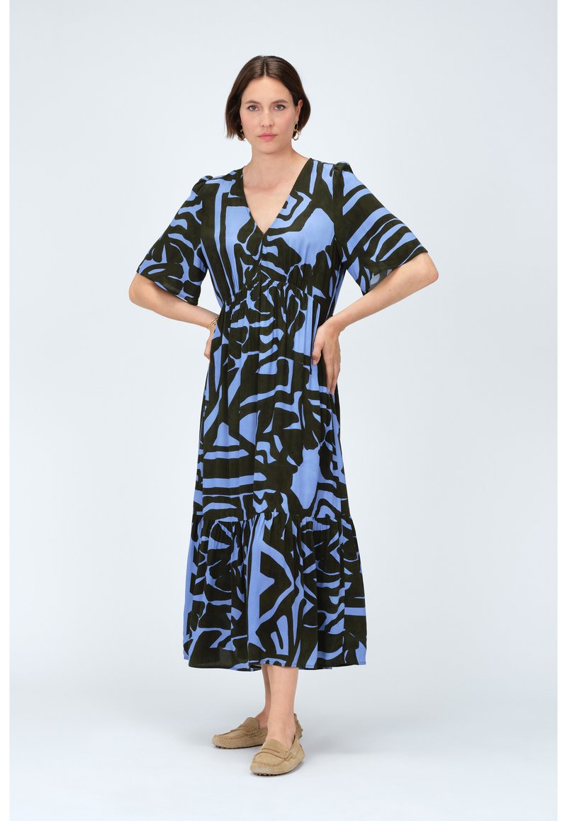 Robe maxi à motifs bleus et noirs avec un décolleté en V, des manches courtes et une jupe à étages, fabriquée en tissu léger. Associée à des chaussures à enfiler beiges.