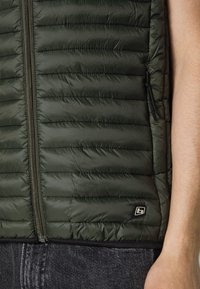 Chaleco acolchado verde con diseño de quilted vertical, cierre de cremallera, bolsillo lateral con cremallera y una pequeña etiqueta de logo cerca del dobladillo. Material de nylon texturizado.