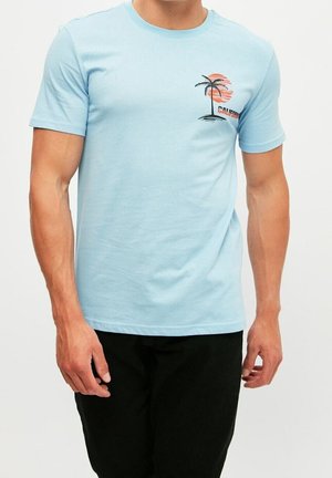 Mann trägt ein hellblaues T-Shirt mit kleinem Palmen- und orangener Sonne-Grafik sowie dem Schriftzug "California", kombiniert mit einer schwarzen Hose.
