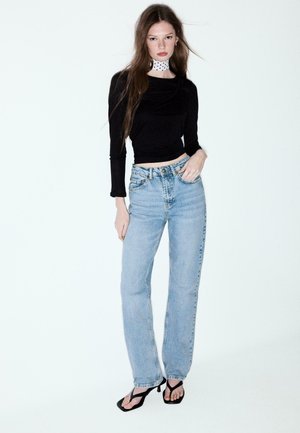 Femme portant un crop top noir à manches longues, un jean taille haute bleu clair, des sandales à talons noires et un foulard blanc à pois autour du cou, debout devant un fond blanc.