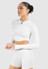 Haut de sport blanc cropped à fermeture éclair et manches longues, texture côtelée, accompagné de shorts taille haute assortis. Présente un design minimaliste et une silhouette ajustée.