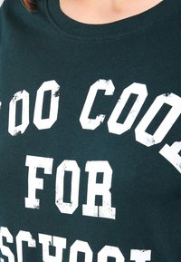 Tmavě zelené bavlněné tričko s velkými bílými blokovými písmeny, které říkají "PŘÍLIŠ COOL NA ŠKOLU." Kolať výstřih, měkká textura, ležérní design.