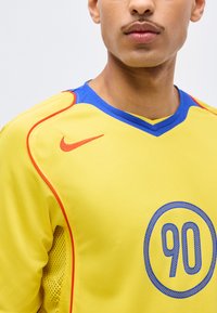 Άνδρας που φοράει κίτρινη αθλητική φανέλα με μπλε γιακά, κόκκινο λογότυπο Nike στον ώμο και μεγάλο μπλε αριθμό 90 τυπωμένο μπροστά.