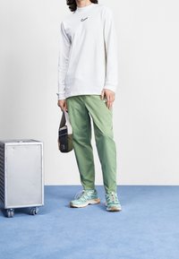 White long-sleeve shirt with black Nike logo; light green pants; mint green sneakers with colorful accents; carrying a small, olive-green bag.