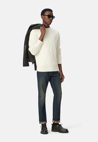 Weißer Pullover mit Kabelstrickmuster, blaue Denim-Jeans, schwarze Lederjacke über der Schulter, schwarze Schuhe und Sonnenbrille. Einfaches, klares Design.