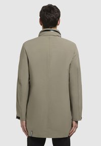 Khaki lang frakke med høj krave, glat tekstur og minimalistisk design. Har sidelæg og sorte accenter ved manchetterne.