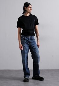 Fekete, rövid ujjú póló zsebbel és vízszintes csíkozással, kék egyenes lábú farmerral, amely kontrasztos varrású és megviselt textúrájú.