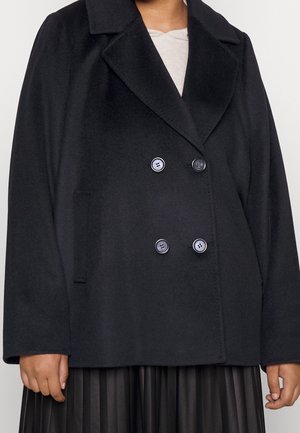 Manteau noir croisé avec larges revers, quatre boutons, poches latérales, porté sur une jupe plissée et un haut clair.