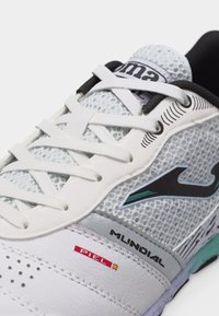 Κοντινό πλάνο λευκού και γκρι αθλητικού παπουτσιού με κορδόνια, επάνω μέρος από δίχτυ, μαύρο λογότυπο και κείμενο «Mundial» και «Piel» στο πλάι.