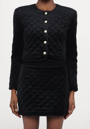 Mujer que lleva una chaqueta corta acolchada de terciopelo negro con mangas largas y botones dorados, y una minifalda acolchada a juego de talle alto.