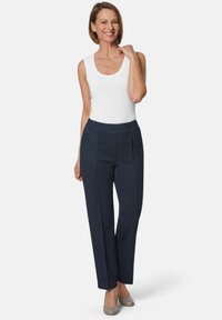 Marineblauwe broek met een ontspannen pasvorm en een elastische tailleband, voorzien van twee plooien aan de voorkant en een gladde textuur. Gecombineerd met een witte tanktop en grijze schoenen.