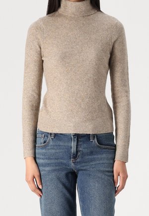 Femme portant un pull à col roulé beige et un jean bleu, debout devant un fond blanc uni.