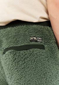 Groene fleece broek met een geribde tailleband, voorzien van een patchzak en een "PRO TEST" logo op een contrasterend label. Zachte, textuurstof.