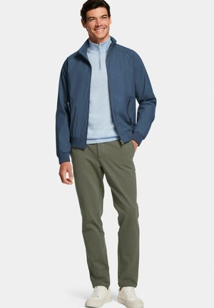 Glunderende man die een blauw jack draagt over een lichtblauwe sweater met rits, olijfgroene broek en witte sneakers, staand met één hand in de zak.