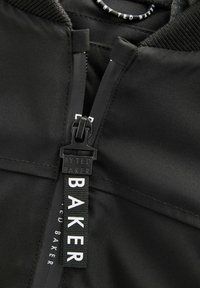 Detailný záber na čiernu bundu s uzáverom na zips a štítkom s názvom značky "By Ted Baker." Tkanina vyzerá hladko a štýlovo.