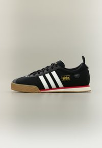 Μαύρο αθλητικό παπούτσι Adidas Samba με λευκές ρίγες, σόλα χρώματος καραμέλας και κόκκινη λεπτομέρεια στη μέση της σόλας, εμφανιζόμενο σε πλευρική προβολή σε ουδέτερο φόντο.