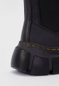 Gros plan sur le talon d’une botte noire avec une semelle extérieure en caoutchouc texturé, des surpiqûres jaunes et des détails en cuir sur la tige, sur fond blanc.