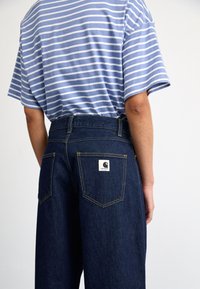 Jean en denim avec une taille haute, couleur bleu foncé, détails de couture jaune, et un patch logo Carhartt sur la poche arrière.