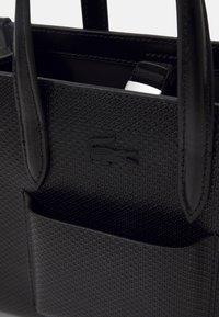Borsa nera con superficie texturizzata, dettagli in pelle, due manici e un logo. Presenta una tasca esterna e un interno spazioso.