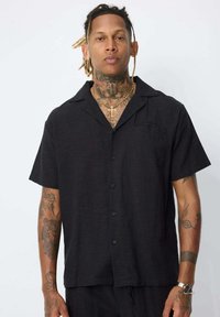 Chemise noire à manches courtes, boutonnée, avec col et tissu texturé, comportant six boutons et un détail de poche avant discret.